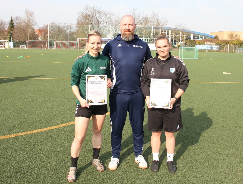 Auf einem Fußballplatz stehen zwei angehende Fußballtrainerinnen mit ihrem Ausbilder. Sie halten Urkunden über ihr Stipendium in den Händen.